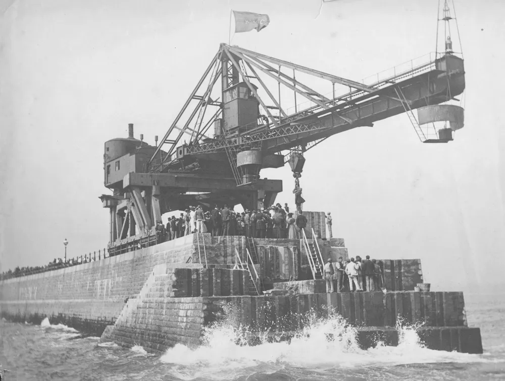 A crowd in hats and long coats stands on the stone end of Roker Pier beneath a huge steel crane, with waves breaking below.